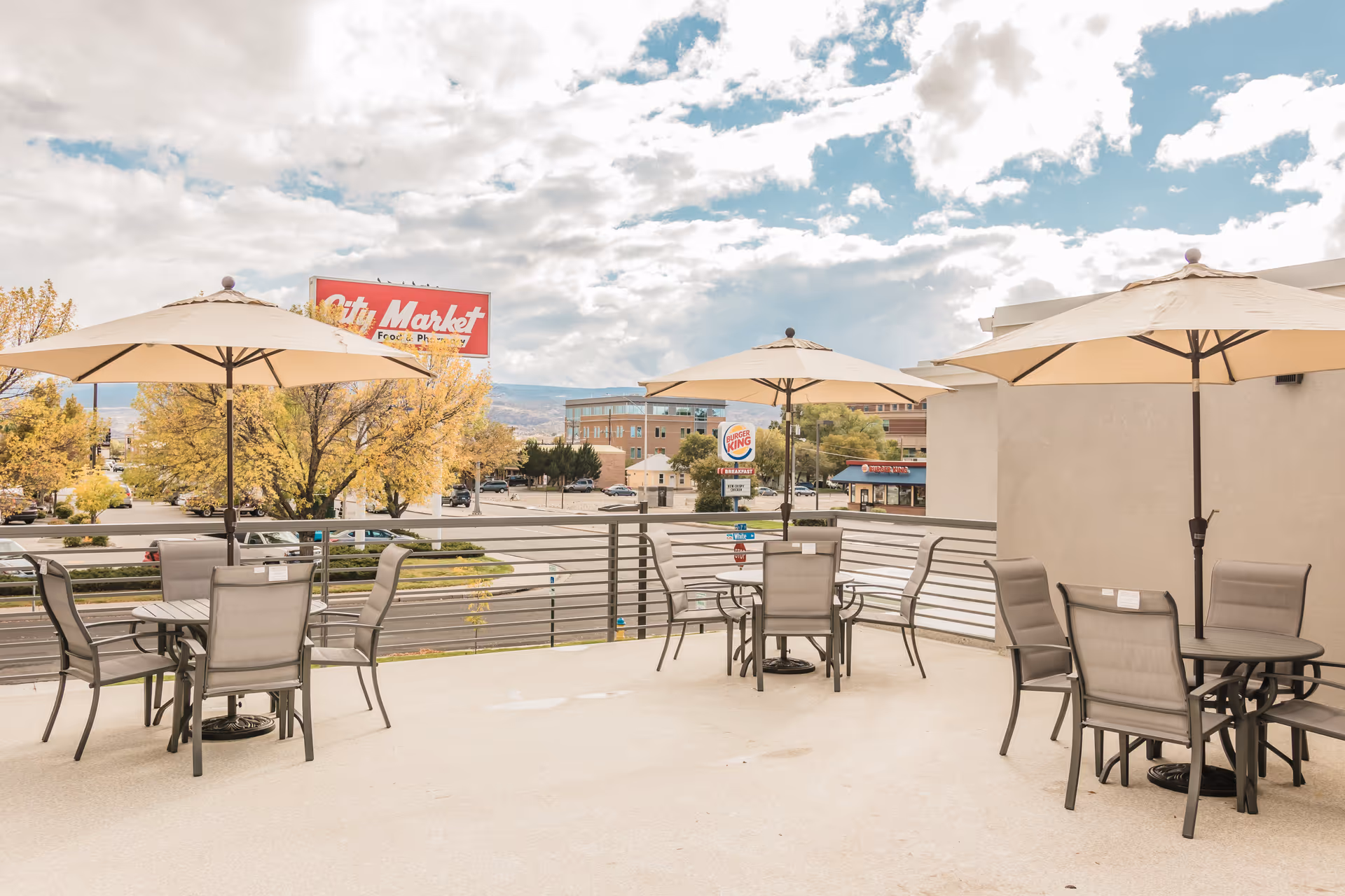Outdoor patio area with three round tables, each surrounded by four chairs and shaded by large beige umbrellas. The patio overlooks a street with visible signs for City Market and Burger King, with trees and buildings in the background under a partly cloudy sky.