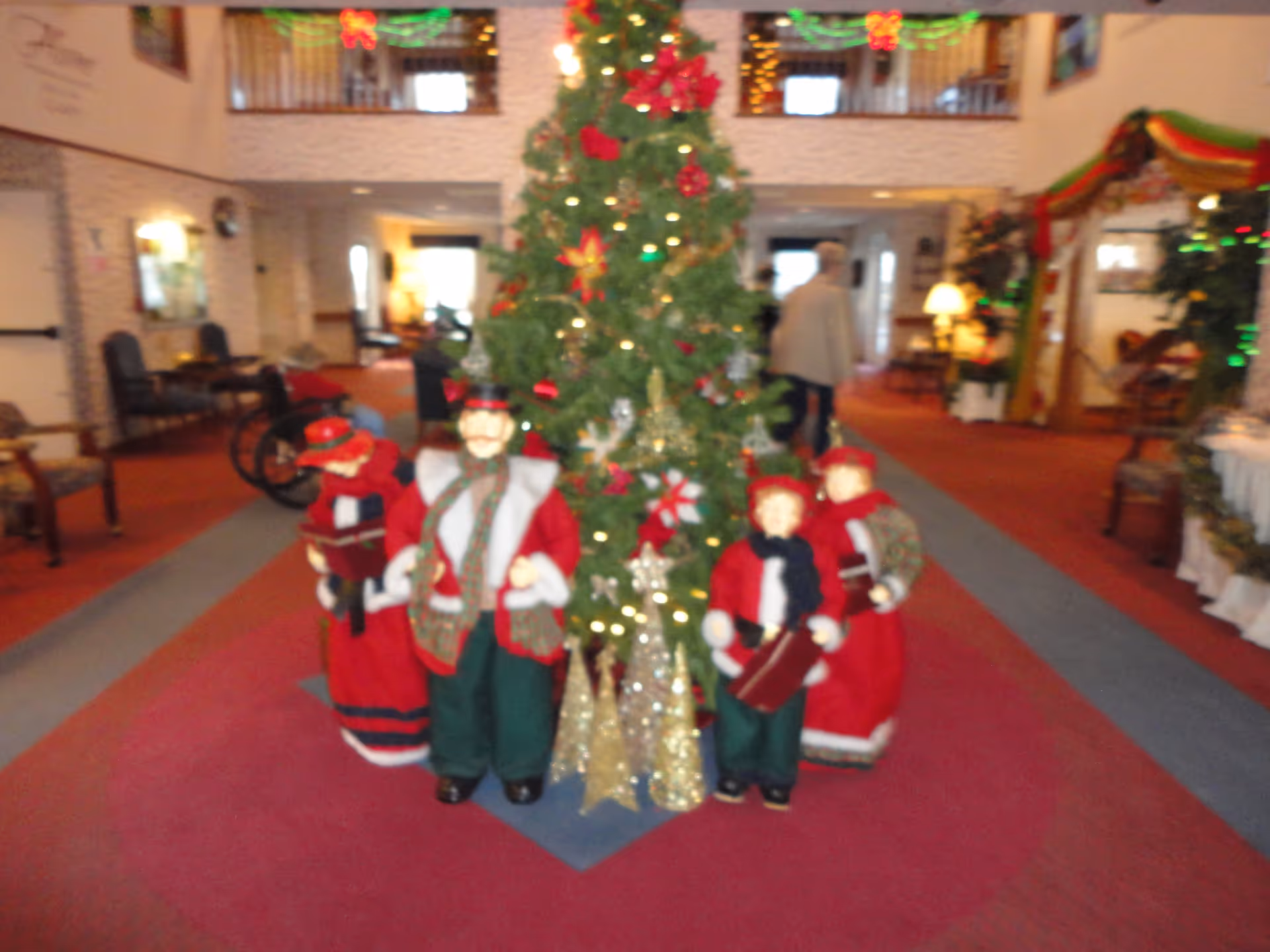 A decorated Christmas tree with lights and ornaments stands in the center of a senior living facility lobby. Around the tree are four festive figurines dressed in red and green holiday clothing. The lobby has red carpeting, seating areas, holiday decorations, and a person walking in the background.
