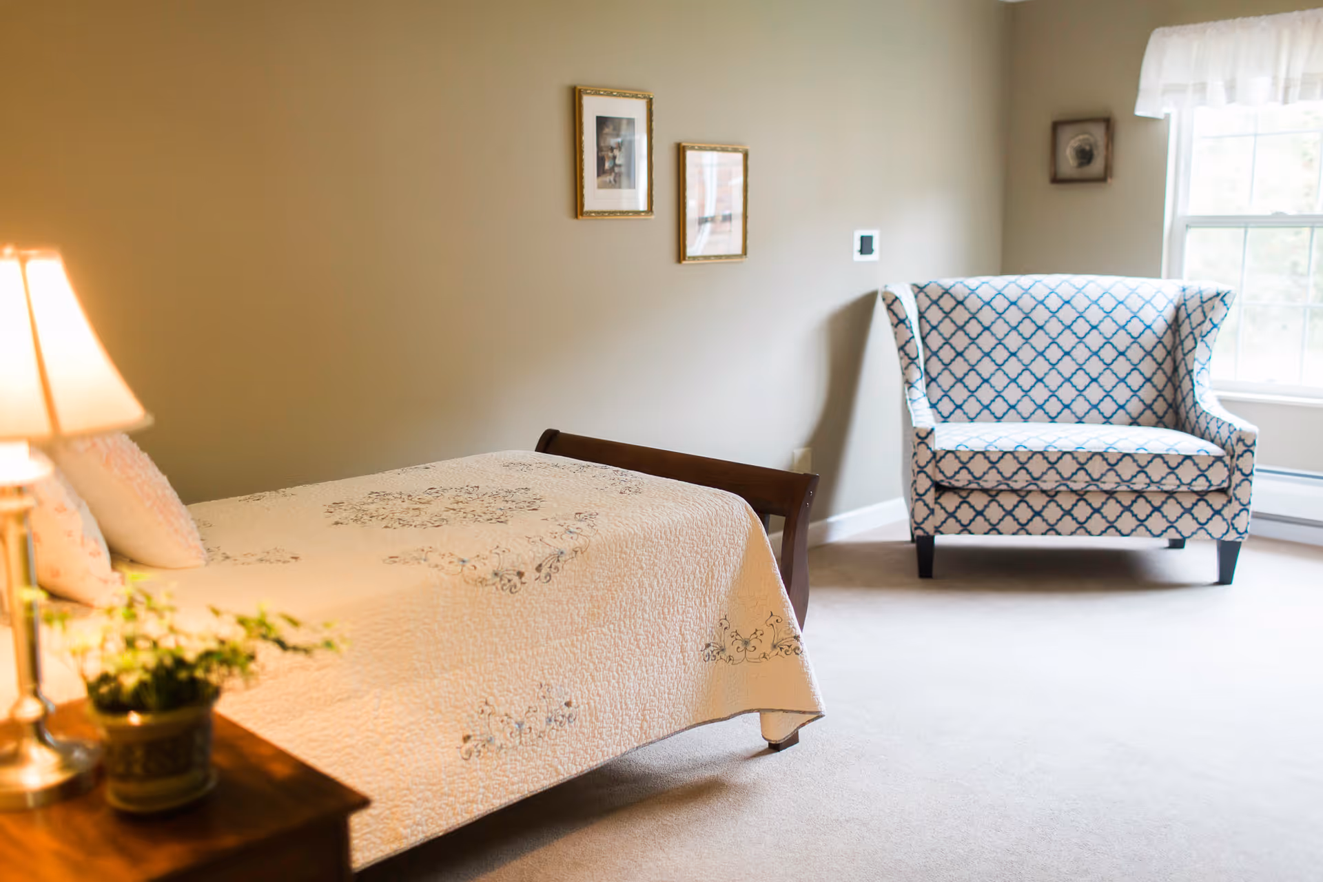 A tidy bedroom with a single bed, a bedside table with a lamp and plant, and a patterned upholstered loveseat by a window.