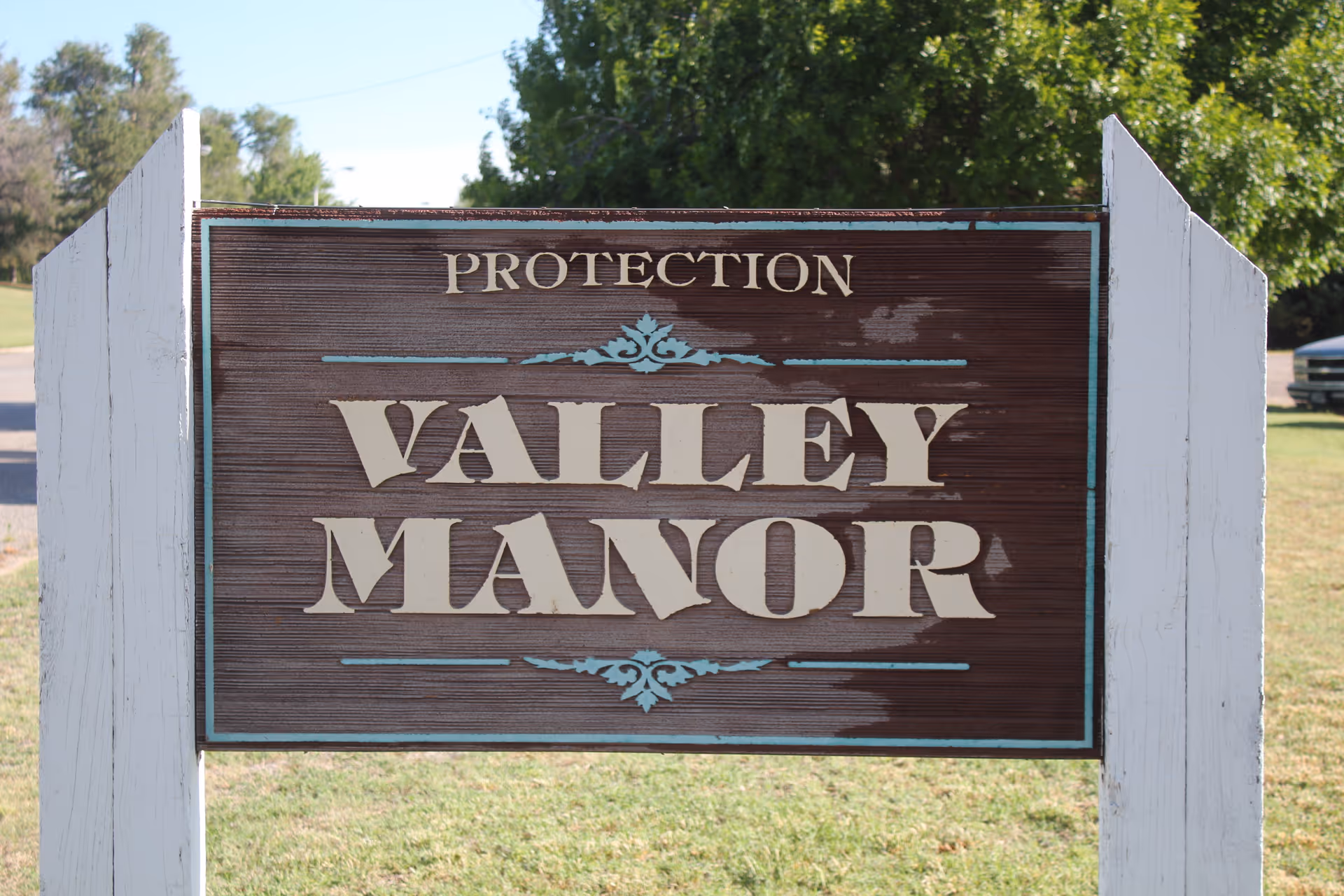 A wooden sign mounted on white posts with the text 'Protection Valley Manor' in large, cream-colored letters with decorative blue accents. The sign is outdoors with grass and trees in the background under a clear sky.