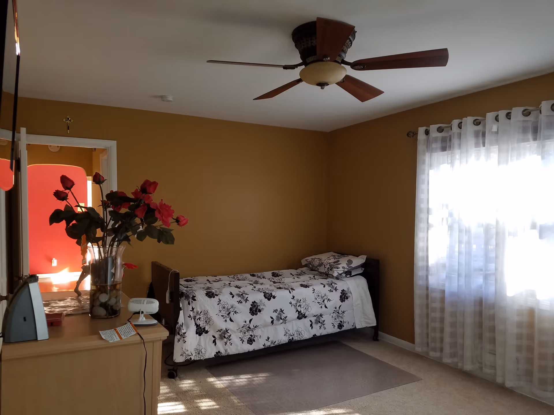 Sunlit bedroom with a single bed in floral bedding, a ceiling fan, sheer curtains, and a dresser topped with a vase of red flowers.