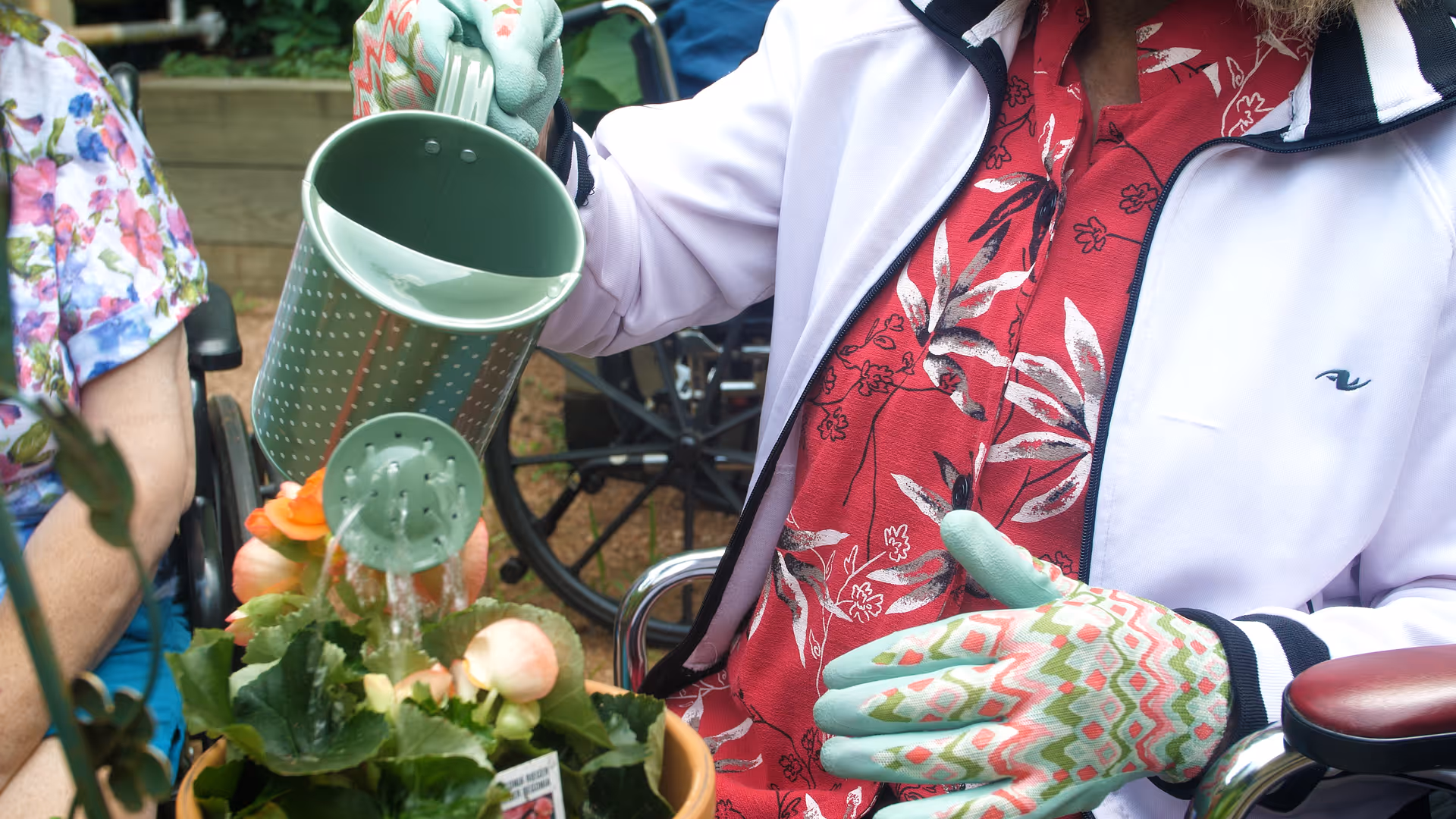 A person wearing patterned gardening gloves and a white jacket with a red floral shirt underneath is watering flowers in a pot using a green watering can. Another person wearing a floral shirt is partially visible sitting nearby in a wheelchair. The scene appears to be outdoors with greenery and wooden planters in the background.