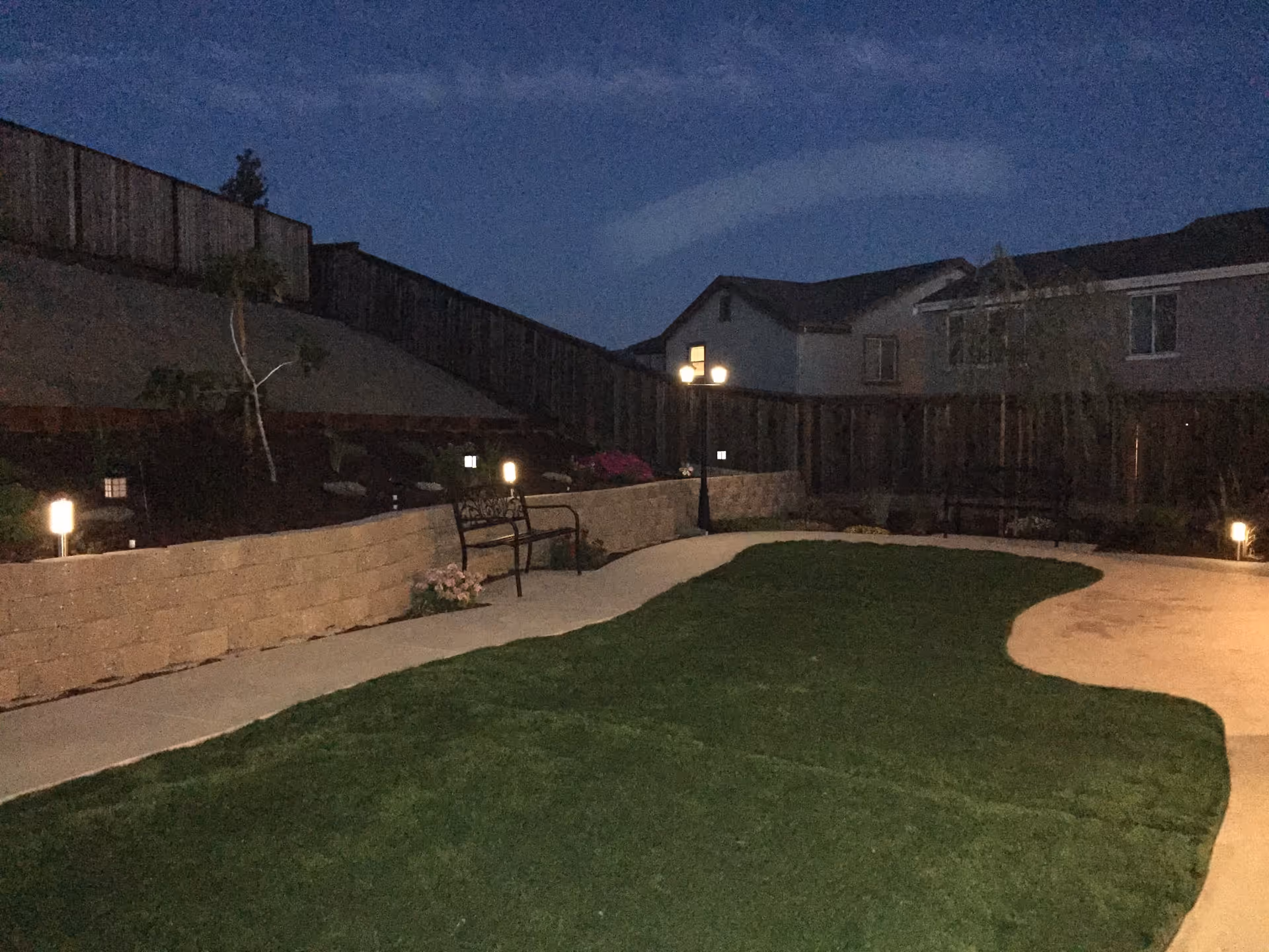 Outdoor garden area at night with a curved concrete pathway, green grass, a low stone retaining wall with small plants, benches, and several small lights illuminating the space. A wooden fence and neighboring houses are visible in the background under a dark sky.