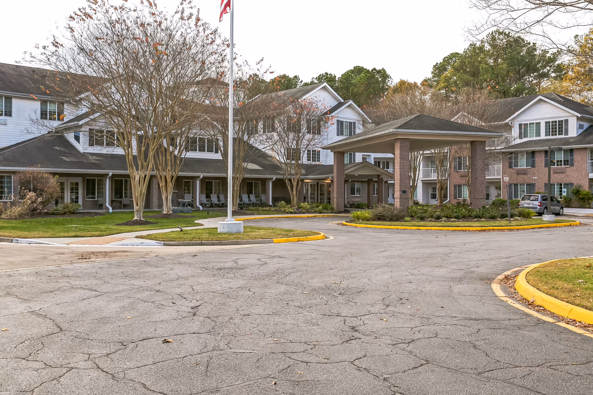 Exterior view of Lighthouse Pointe by Barclay House, showing a multi-story senior living facility with a covered entrance, an American flag on a flagpole, leafless trees, and a paved driveway with yellow curbs.