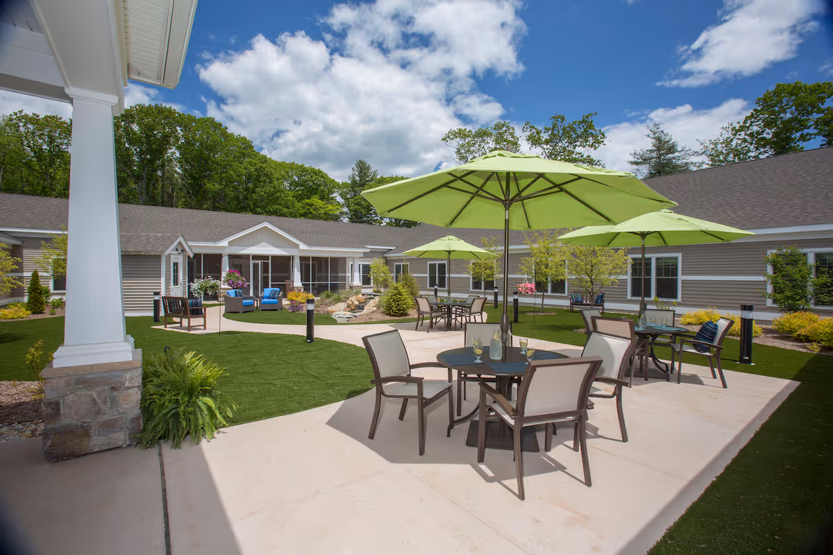 Outdoor patio area at Cornerstone at Hampton with round tables and chairs under green umbrellas, surrounded by a well-maintained lawn and landscaping, with a building in the background under a partly cloudy blue sky.