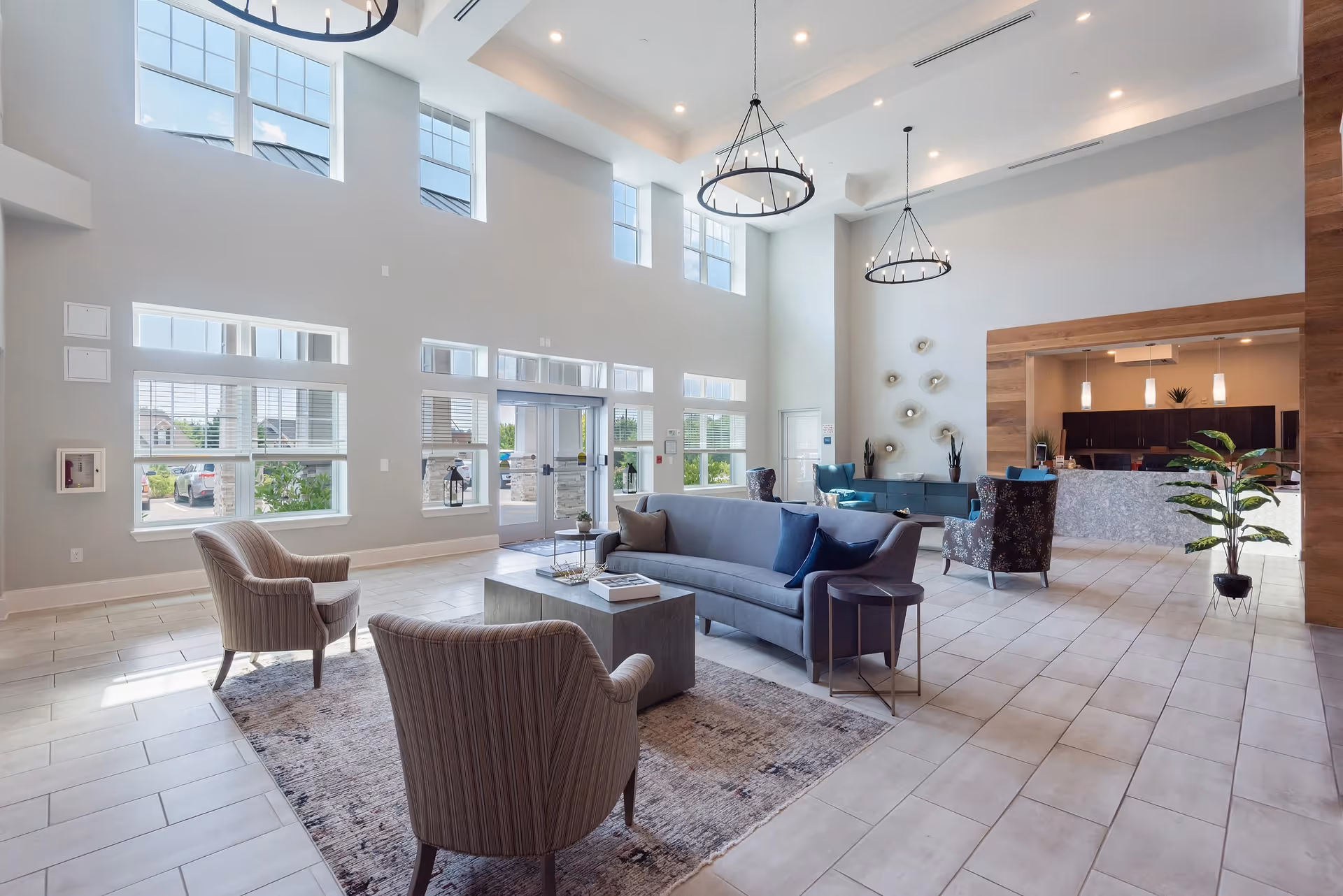 Bright and spacious senior living facility lobby with high ceilings and large windows letting in natural light. The seating area includes a gray sofa, two striped armchairs, a coffee table on a patterned rug, and additional chairs near a reception desk. Modern chandeliers hang from the ceiling, and there are decorative plants and wall art.