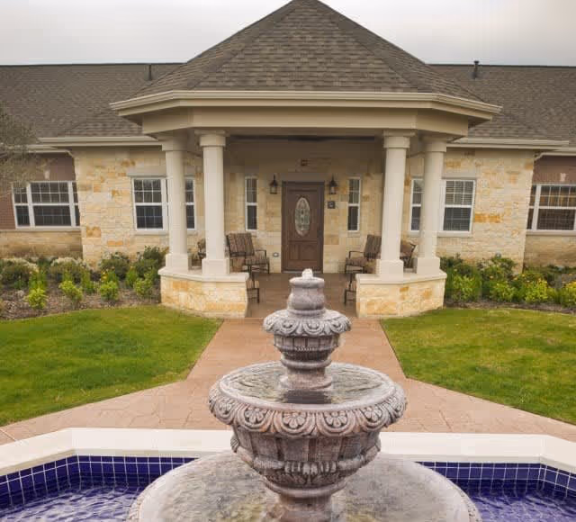 Front entrance of a building with a covered porch supported by four columns, two chairs on the porch, surrounded by landscaped greenery and a stone fountain in the foreground.