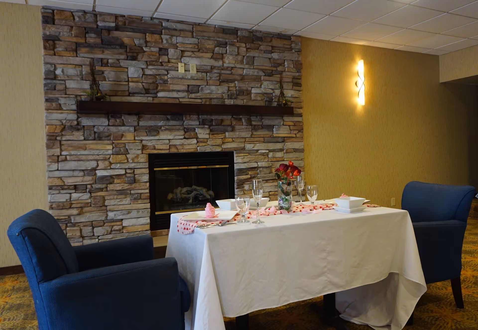 Table set for two with a white tablecloth, glassware and a vase of flowers between two blue armchairs in front of a stone fireplace in a lounge area.