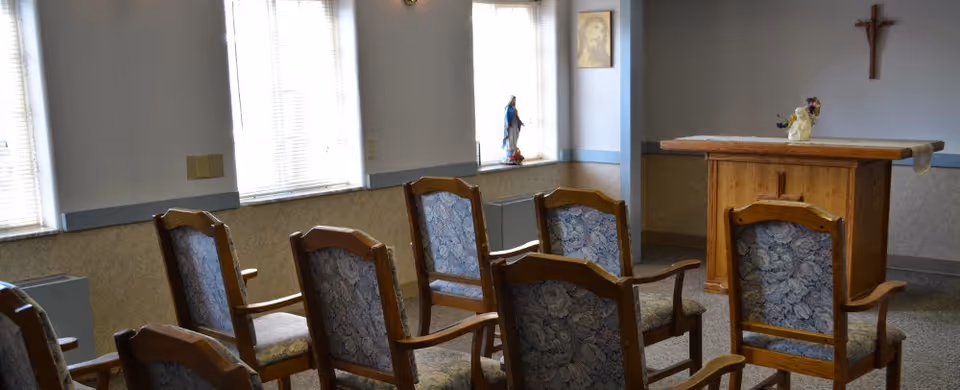 A small chapel or prayer room with wooden chairs arranged in rows facing a wooden altar. The room has windows with blinds, religious statues on the windowsills, a framed picture on the wall, and a crucifix mounted above the altar.