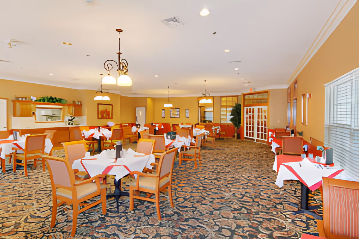 Spacious dining room with multiple round and rectangular tables set with white and red tablecloths, wooden chairs, chandeliers, and patterned carpet.