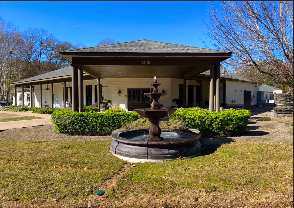 Exterior view of a single-story building with a covered entrance supported by columns, a three-tiered water fountain in the foreground, green bushes, and leafless trees under a clear blue sky.