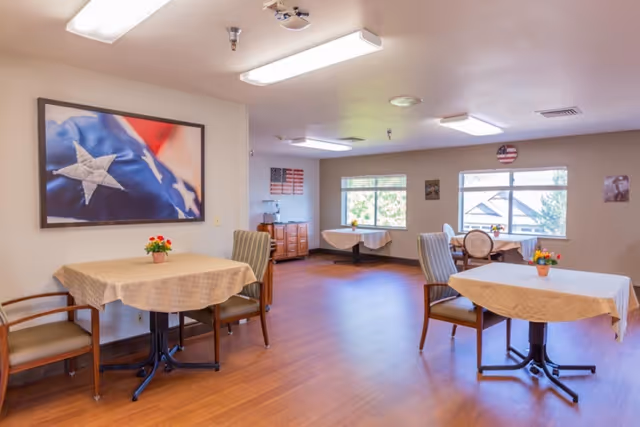 A bright dining room with several tables covered with beige tablecloths and small flower pots as centerpieces. The room has wooden flooring, multiple chairs around the tables, and large windows letting in natural light. On the walls are patriotic-themed decorations including a large framed photo of a folded American flag and smaller American flag artwork.