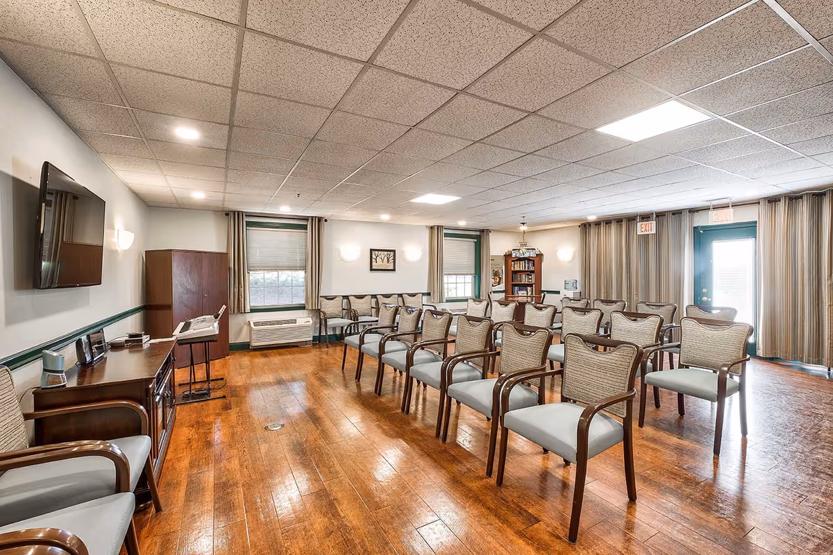 A well-lit meeting or activity room with rows of cushioned chairs arranged facing a wall-mounted flat screen TV. The room has wooden floors, two windows with blinds and curtains, a wooden cabinet, a bookshelf, and an exit door with curtains. The ceiling has recessed lighting and acoustic tiles.