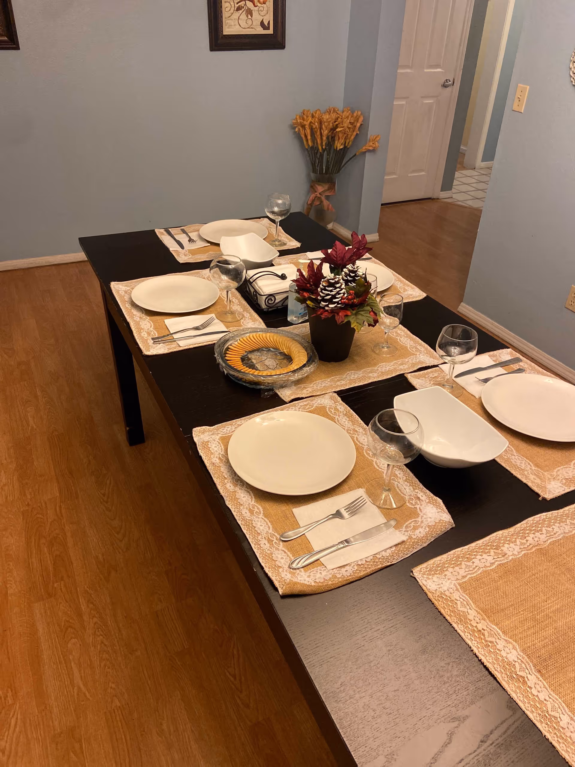 A dining table set for six with placemats, plates, glassware and a floral centerpiece in a home dining room.