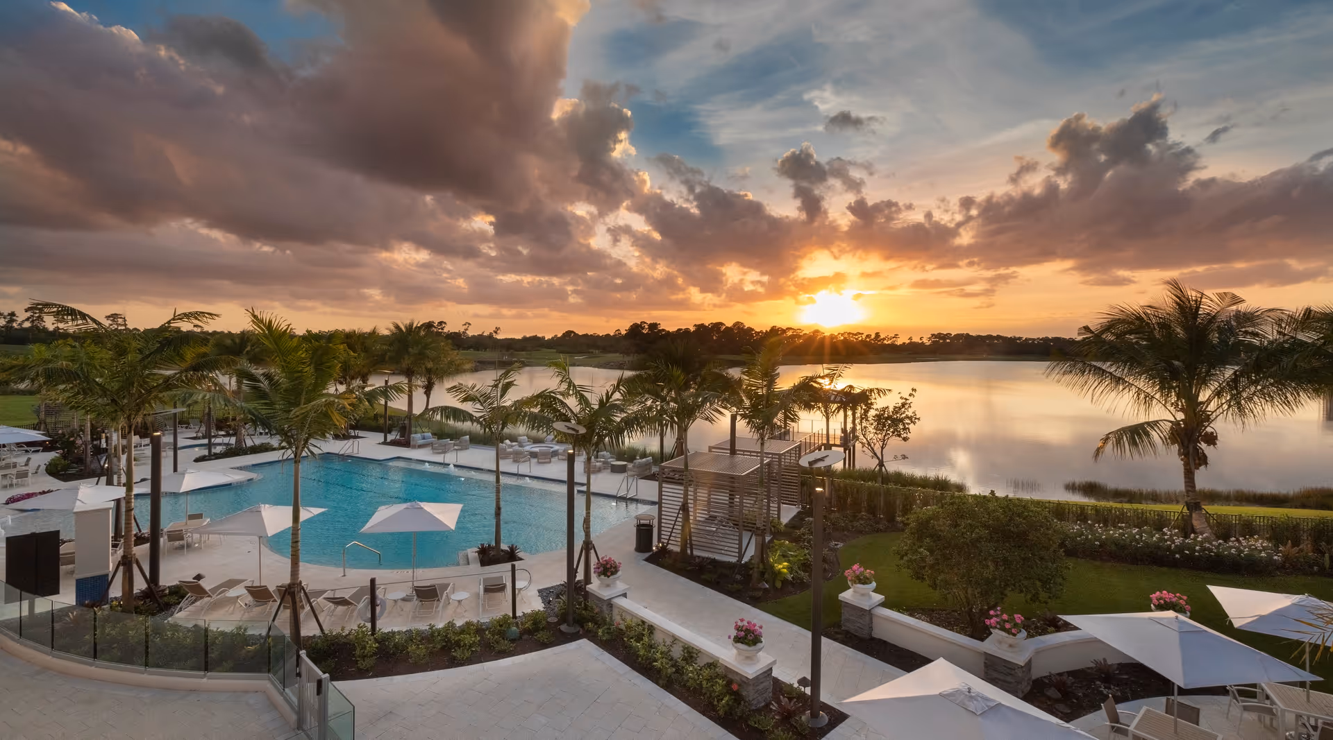 Outdoor view of a swimming pool area with lounge chairs and umbrellas surrounded by palm trees, overlooking a large lake at sunset with dramatic clouds in the sky.