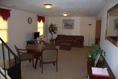 Interior view of a living room area in an assisted living facility featuring a round wooden table with four chairs, a brown sofa with two ottomans, a floor lamp, a potted plant, a wall clock, framed artwork, and a large mirror on the wall. The room has beige carpeting and two windows with red valances.