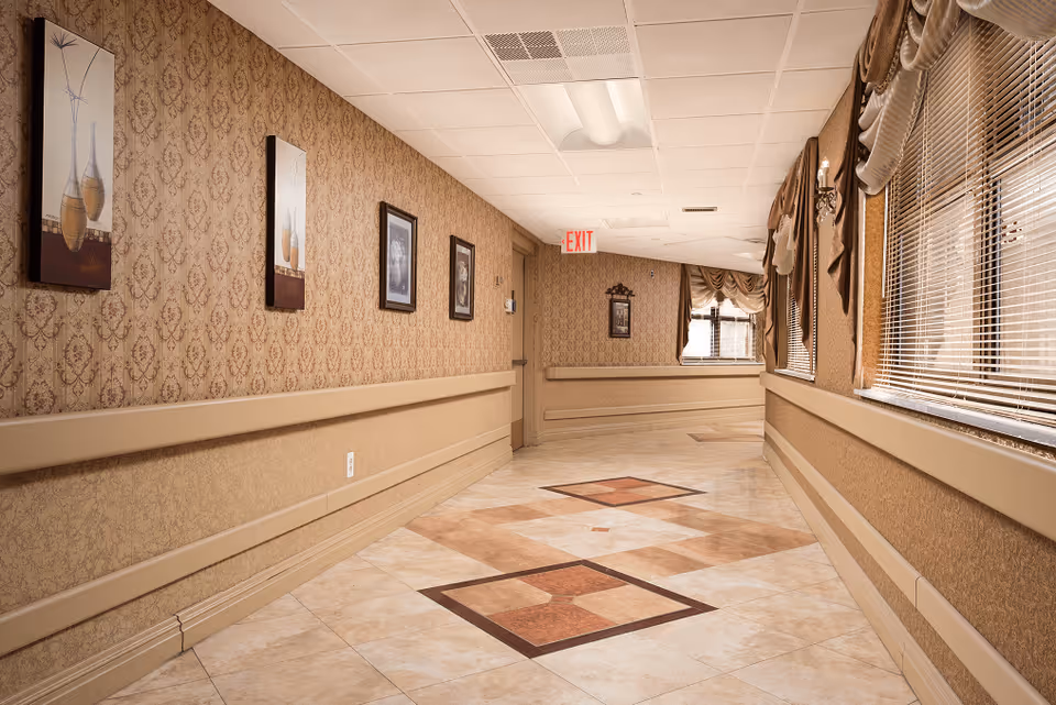 A well-lit hallway in a senior living facility with patterned wallpaper, framed artwork on the left wall, large windows with blinds and draped curtains on the right, and decorative tile flooring. An exit sign is visible at the end of the hallway.