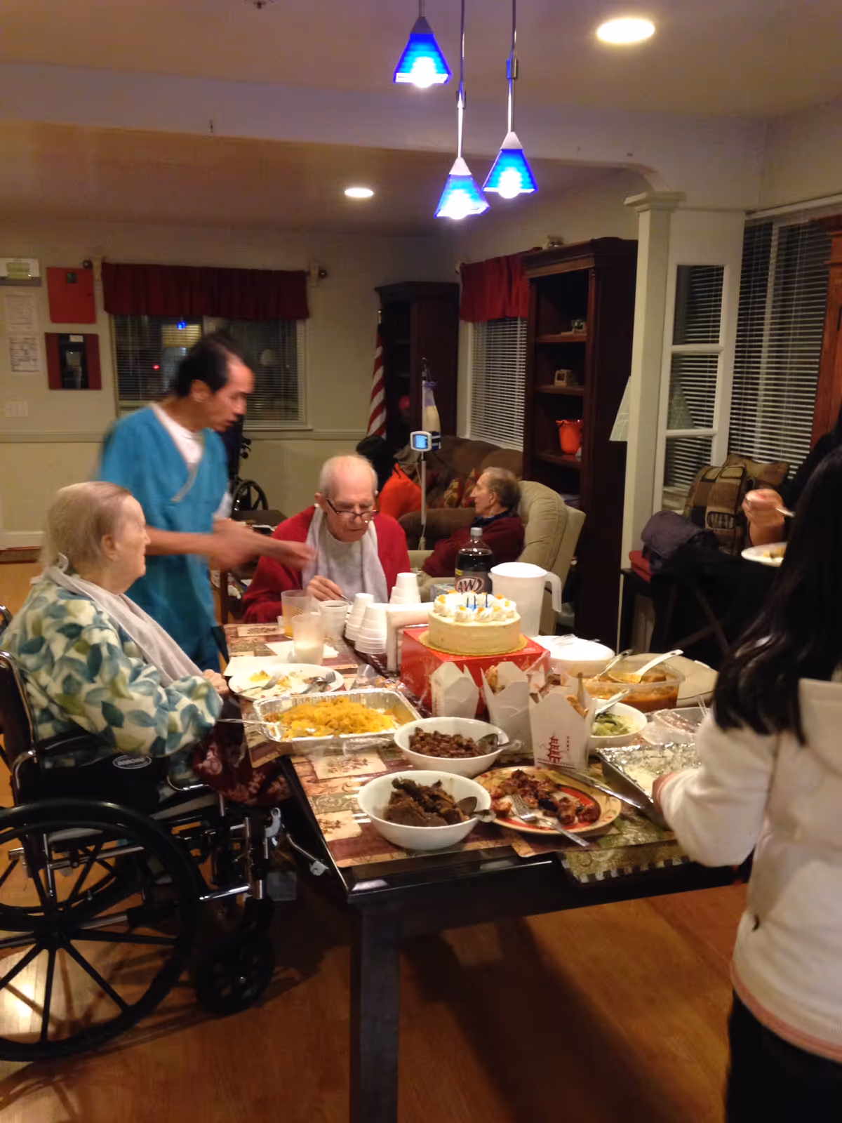 A group of elderly people and a caregiver gathered around a dining table with various dishes and a birthday cake. One elderly person is in a wheelchair, and the caregiver is serving or assisting. The setting appears to be a cozy common area with bookshelves, windows with blinds, and warm lighting.