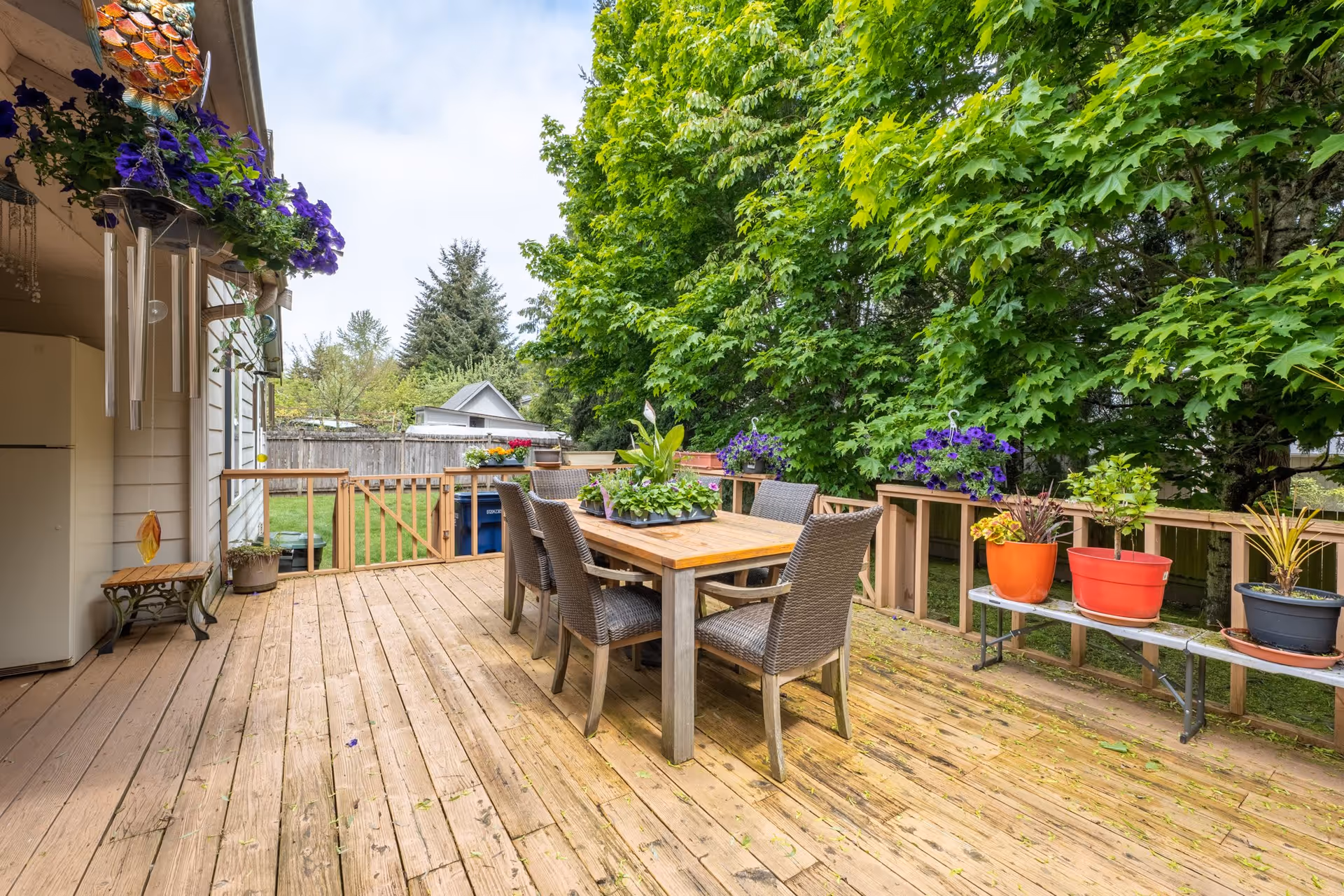 Wooden outdoor deck with a dining table and six wicker chairs surrounded by potted plants and hanging flower baskets, with a large green tree and a fenced backyard in the background.