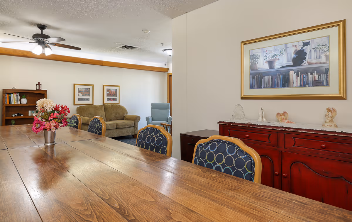 A communal dining room with a long wooden table and chairs, a vase of flowers, a red sideboard, and sofas in the background.