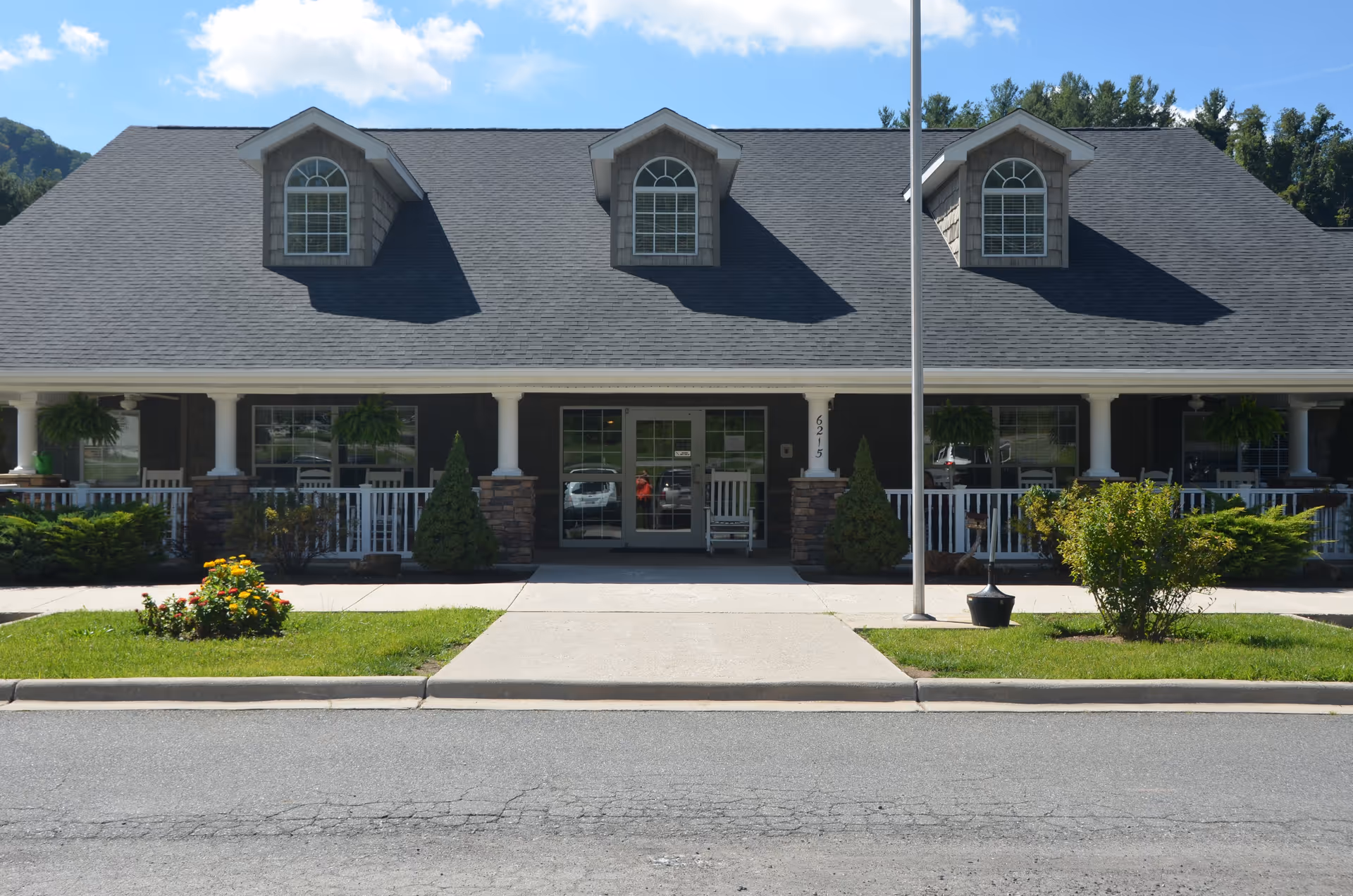 Front exterior view of a single-story building with a dark shingled roof featuring three dormer windows. The building has a covered porch with white columns and railings, stone accents on the lower walls, and a central entrance with double glass doors. There is a concrete walkway leading to the entrance, surrounded by green grass, bushes, and flowers. A flagpole is positioned near the walkway.