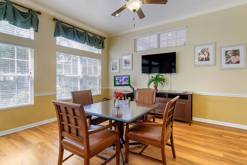 Sunlit dining room with a wooden table and four chairs, large windows with green valances, a wall-mounted TV, and a sideboard with a plant.
