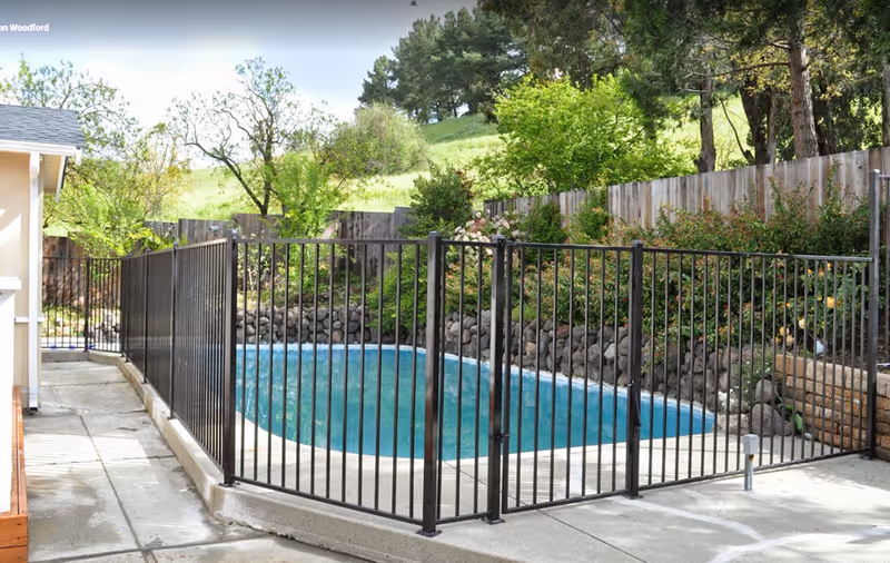 Outdoor swimming pool enclosed by a black metal fence with a gate, surrounded by greenery, trees, and a wooden fence in the background, with a concrete walkway beside the pool area.