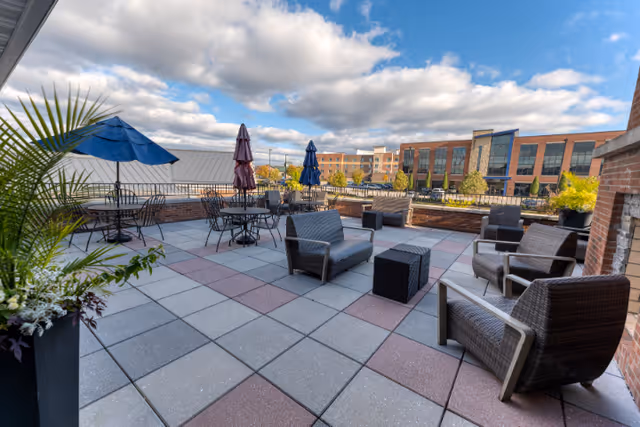 Outdoor patio area with multiple seating arrangements including wicker chairs, sofas, and tables with umbrellas under a partly cloudy sky. The patio is paved with square tiles in shades of gray and red, surrounded by brick walls and planters with greenery. In the background, there are modern buildings and trees.