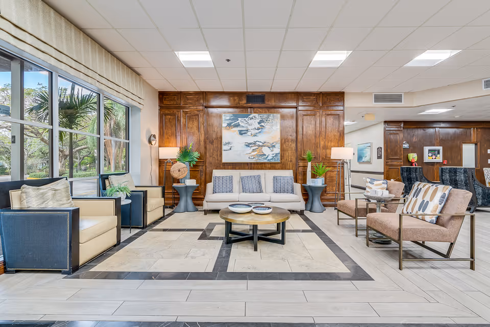 A bright and spacious senior living facility lounge area with large windows on the left showing greenery outside. The room features a beige sofa with three blue cushions, two beige armchairs with patterned pillows, two black and beige armchairs, and round coffee tables in the center. The back wall is wooden paneled with a large abstract painting and two floor lamps with plants on side tables.