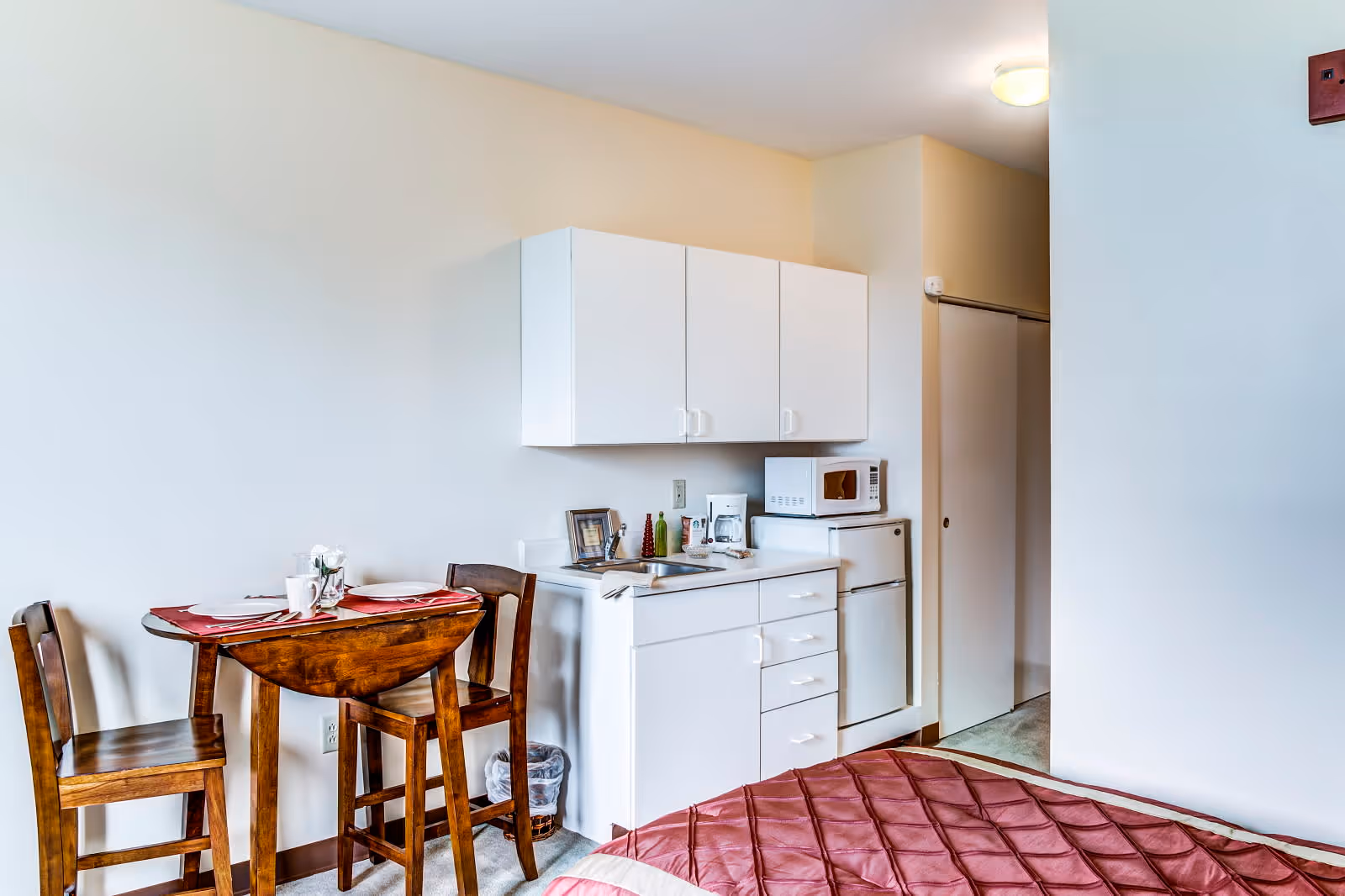 A small studio apartment interior featuring a compact kitchenette with white cabinets, a microwave, a mini refrigerator, and a coffee maker. Next to the kitchenette is a small wooden dining table with two chairs, set with placemats, plates, and cups. Part of a bed with a red quilted bedspread is visible in the foreground.