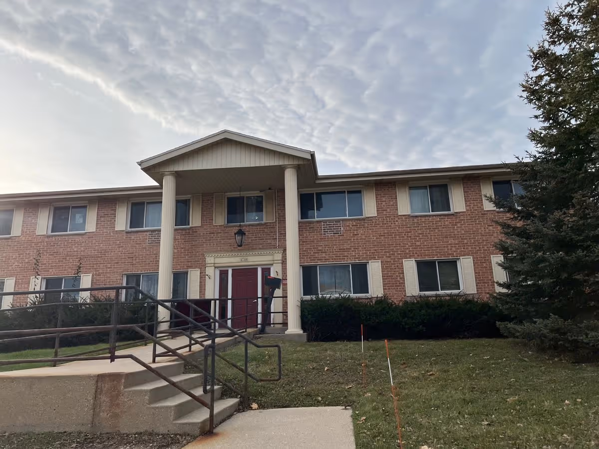 Two-story brick senior living building with columns, a central red entrance, steps and a ramped walkway.