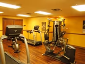 Interior view of a senior living facility fitness room with exercise equipment including treadmills and a multi-function weight machine, wooden flooring, and warm yellow walls with ceiling lights.