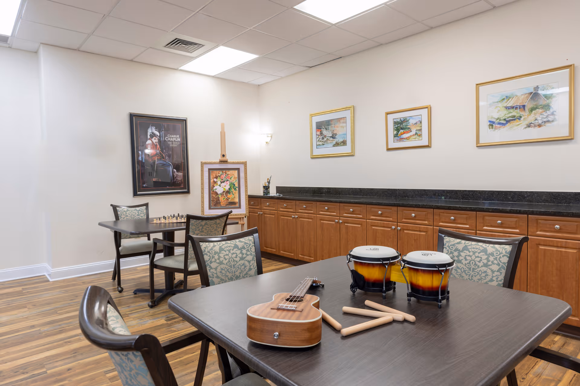 A well-lit room with wooden flooring and light-colored walls featuring framed artwork. There are two tables with cushioned chairs around them. On the table in the foreground, there is a ukulele, two bongo drums, and drumsticks. The back wall has a long counter with wooden cabinets underneath.
