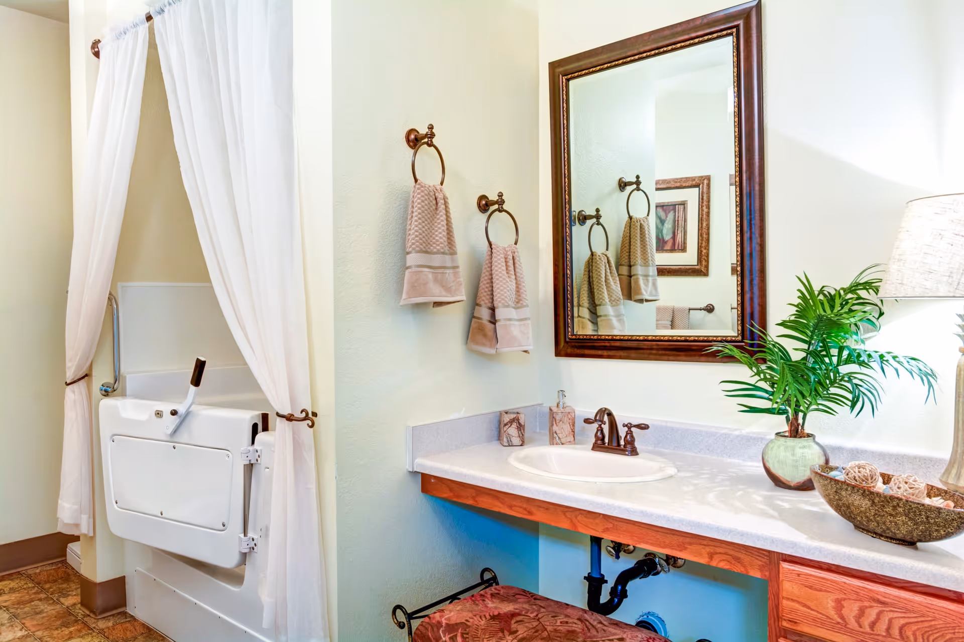 A bathroom with a white walk-in bathtub partially covered by white curtains. There is a vanity with a sink, a large framed mirror above it, two beige hand towels hanging on bronze towel rings, a potted plant, a lamp, and decorative items on the countertop. The floor has a brown tile pattern.
