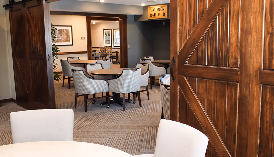 Interior view of a senior living facility dining area with several round tables and cushioned chairs. The room features wooden sliding barn doors and a sign above the doorway that reads 'WOODEN RAIL PUB'. The space is carpeted and decorated with framed artwork on the walls.