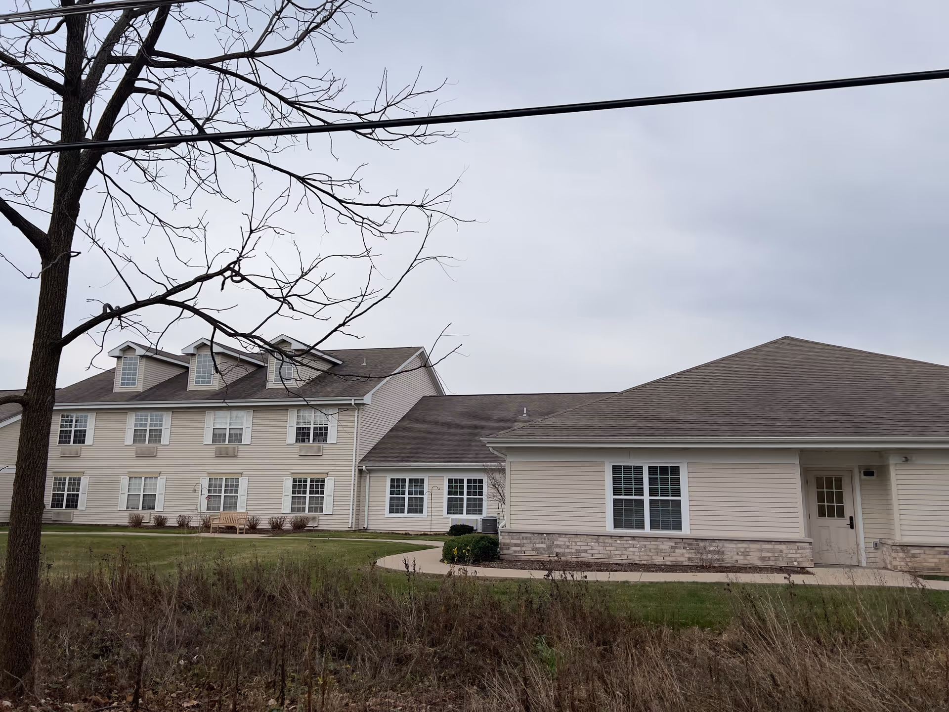 Exterior view of a beige senior living facility building with multiple windows and a sloped roof under a cloudy sky. There is a leafless tree in the foreground and a grassy area with a sidewalk leading to the entrance.