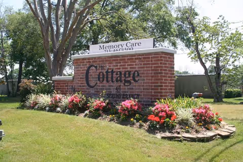 Brick entrance sign for 'The Cottage of Spring Branch' surrounded by landscaped flowers and trees.