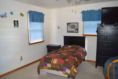 Small bedroom with a single bed, dresser, two windows, blue valances, and a ceiling fan.