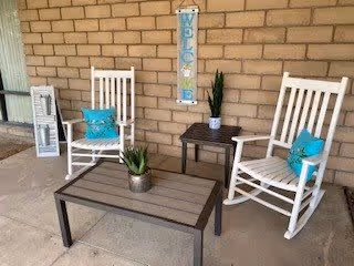 A cozy outdoor seating area with two white rocking chairs each with a turquoise pillow, a small dark wooden side table with a potted plant, and a larger matching coffee table with another potted plant. The background features a brick wall with a vertical 'WELCOME' sign hanging on it.
