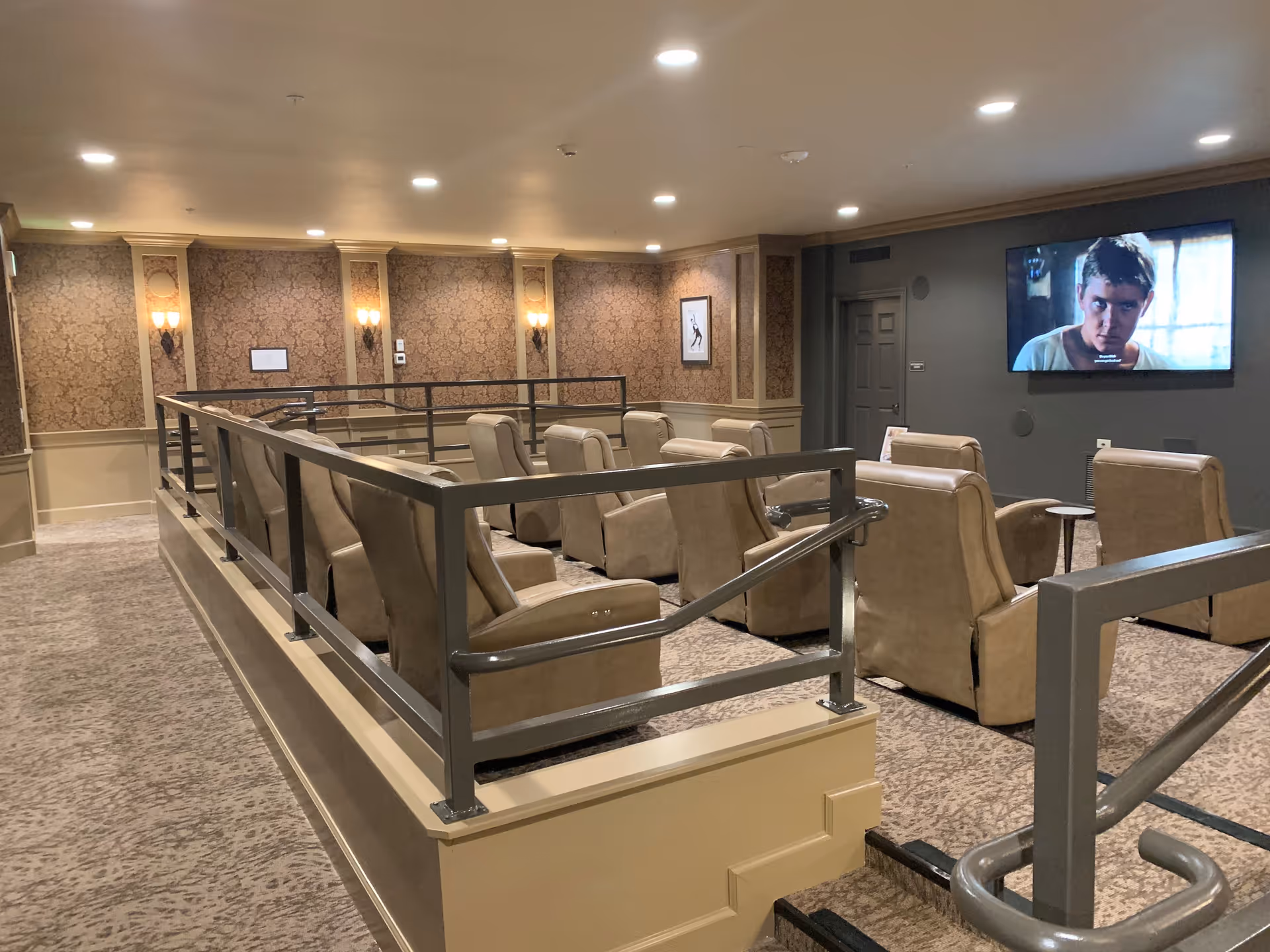 Interior view of a senior living facility's media room with multiple beige recliner chairs arranged in rows facing a wall-mounted flat screen TV. The room has patterned wallpaper, wall sconces, and recessed ceiling lights, with a metal railing surrounding the seating area.