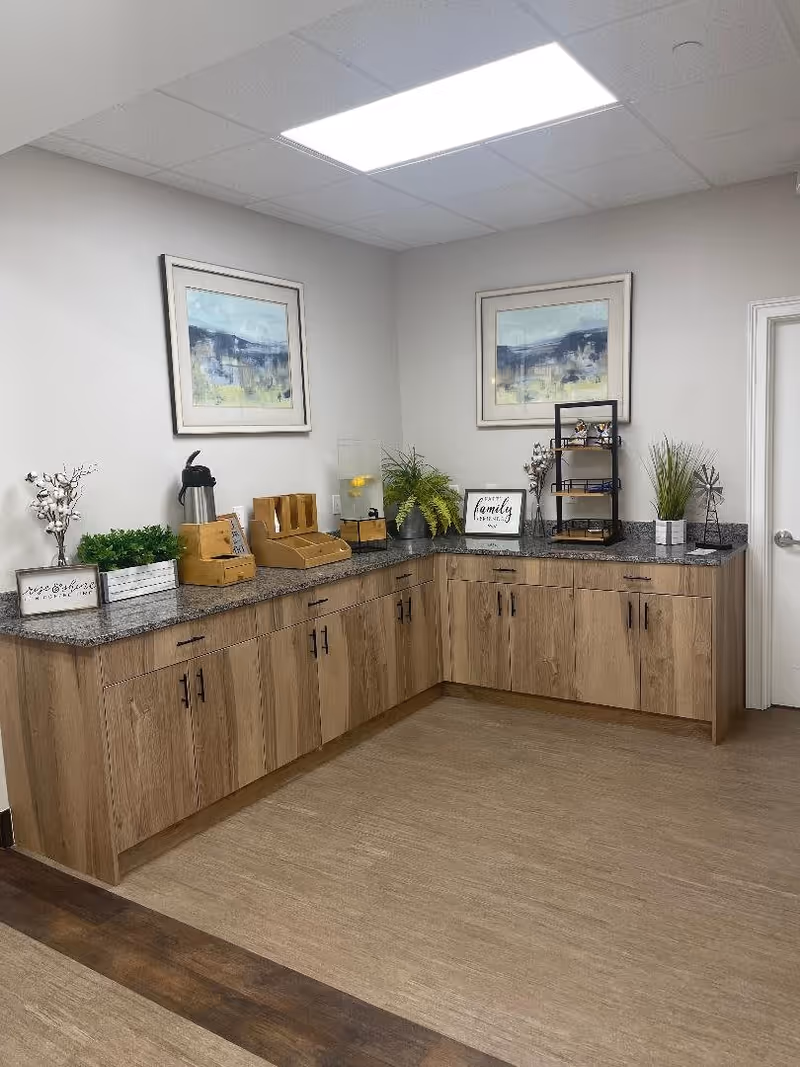 Corner countertop area with wooden cabinets below and granite surface above, decorated with plants, framed artwork, a coffee dispenser, and small decorative items in a well-lit room with light-colored walls and ceiling tiles.