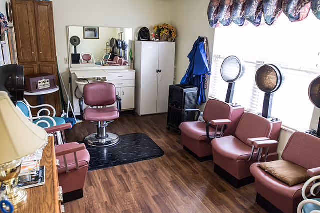 Interior of a salon room with wooden flooring, featuring three maroon salon chairs with attached hair dryers near a window with blinds and a valance. There is a large mirror above a white cabinet with drawers, a single salon chair in front of it, and various salon equipment and furniture including a lamp, a tall wooden cabinet, and a white storage cabinet.
