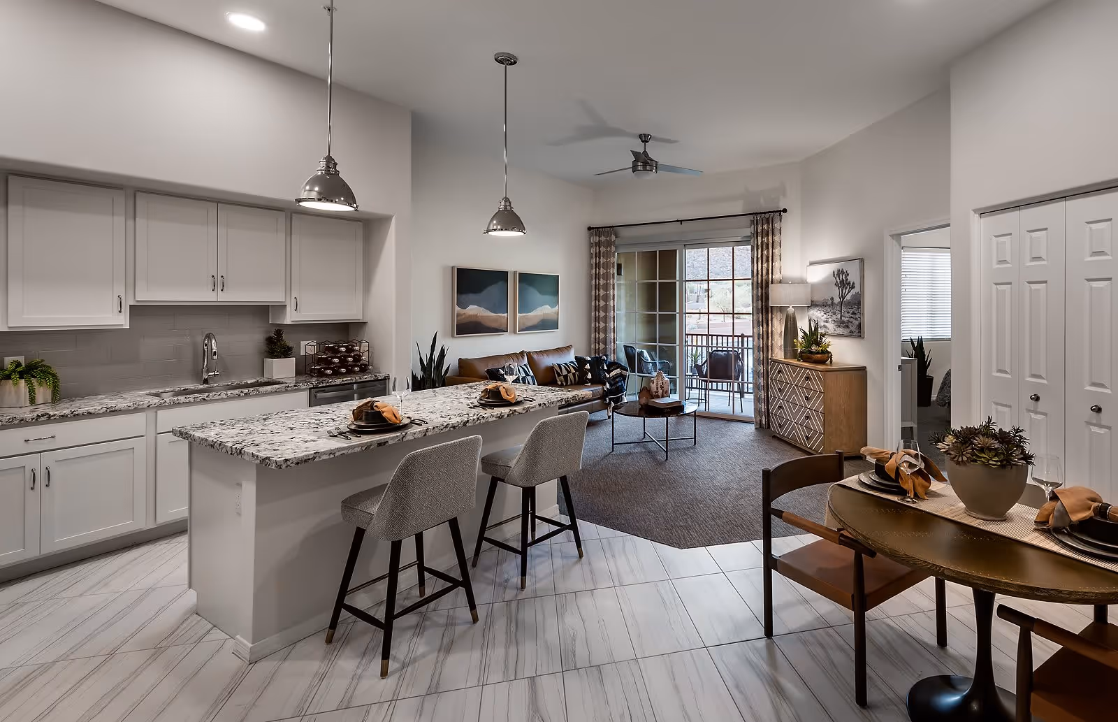 Modern open-concept living space featuring a kitchen with white cabinets, granite countertops, and two pendant lights above a breakfast bar with two upholstered stools. Adjacent to the kitchen is a cozy living room with a brown sofa, coffee table, and sliding glass doors leading to a balcony. A small dining area with a round table and four chairs is also visible, along with decorative plants and artwork on the walls.