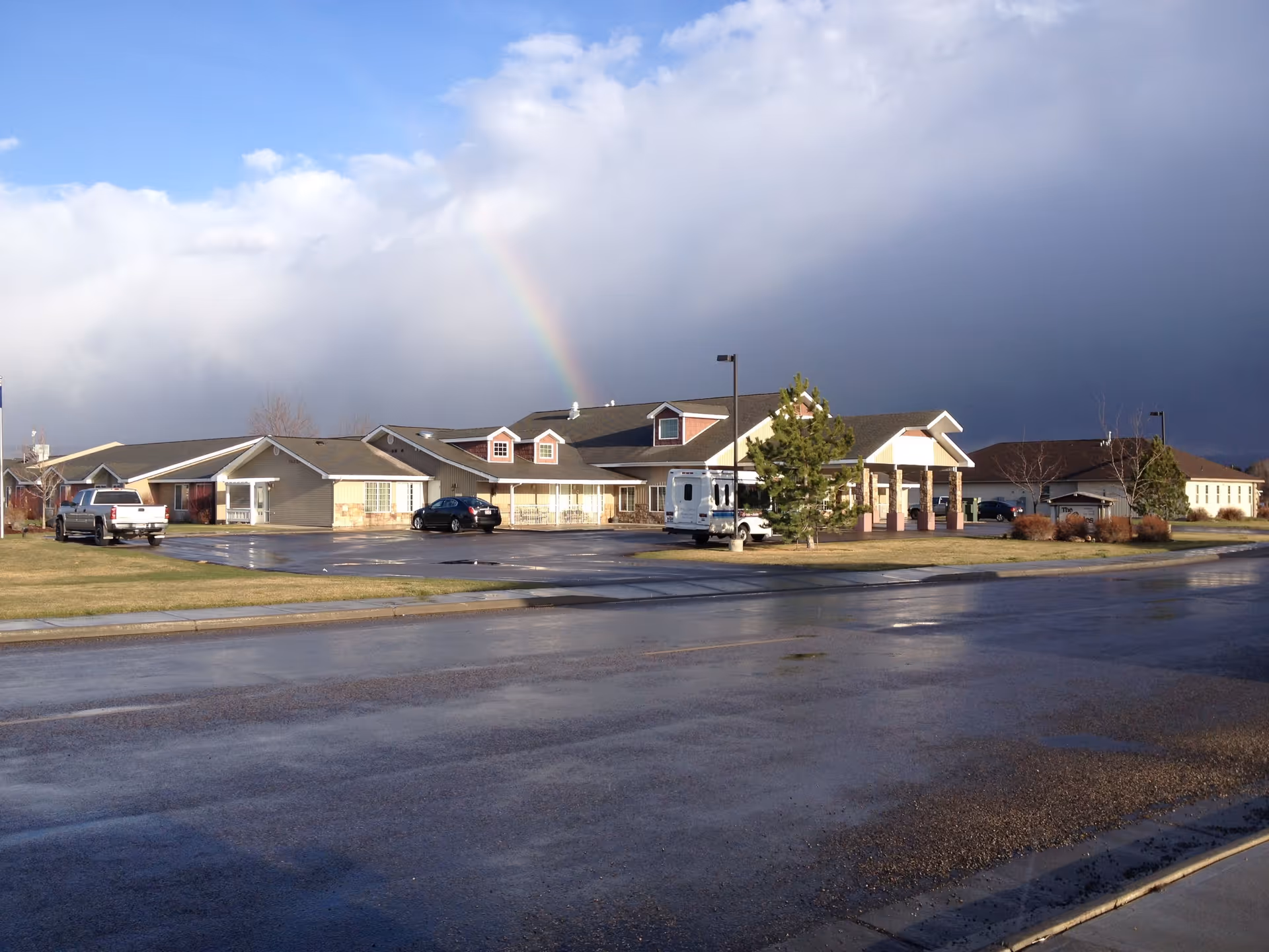 Front exterior of a single-story assisted living facility with parked vehicles, wet pavement, and a rainbow in the sky.
