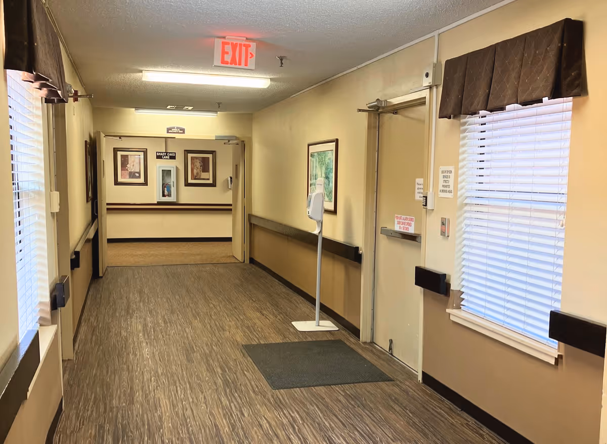 A well-lit senior living hallway with wood-look flooring, handrails, framed artwork, windows with blinds, and an illuminated EXIT sign.