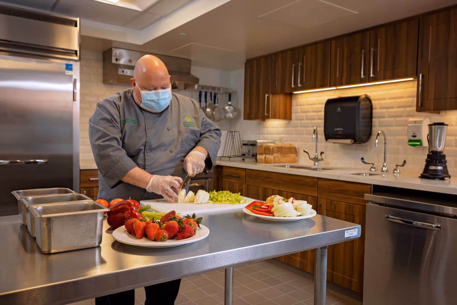 A chef wearing a face mask and gloves is slicing onions on a cutting board in a clean, modern kitchen. The kitchen has wooden cabinets, stainless steel appliances, and plates of fresh fruits and vegetables on a metal countertop.