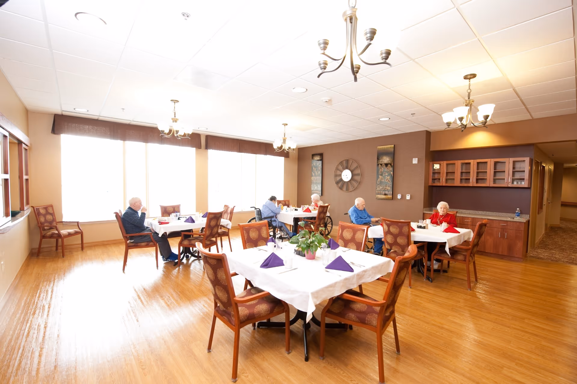 Bright dining room with several elderly residents seated at tables set with white tablecloths and purple napkins.