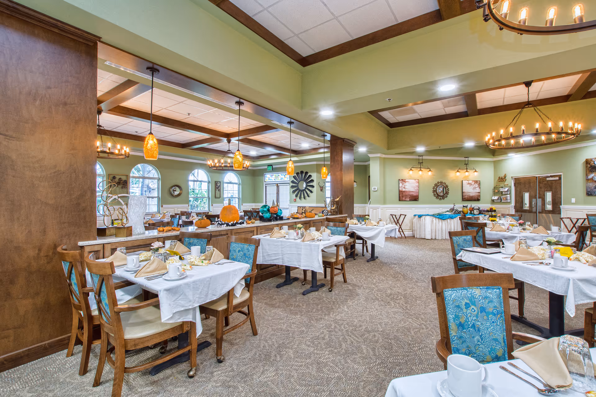 A spacious dining room in an assisted living facility with multiple tables set with white tablecloths, beige napkins, cups, and silverware. The room features green walls, large windows, decorative lighting fixtures including chandeliers and pendant lights, and autumn-themed decorations such as pumpkins on a central counter.