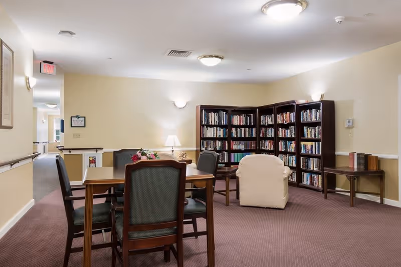 A cozy reading and sitting area in a senior living facility featuring a wooden table with four chairs, a beige armchair, a lamp on a side table, and multiple bookshelves filled with books against the wall. The room has beige walls, carpeted floor, and ceiling lights.