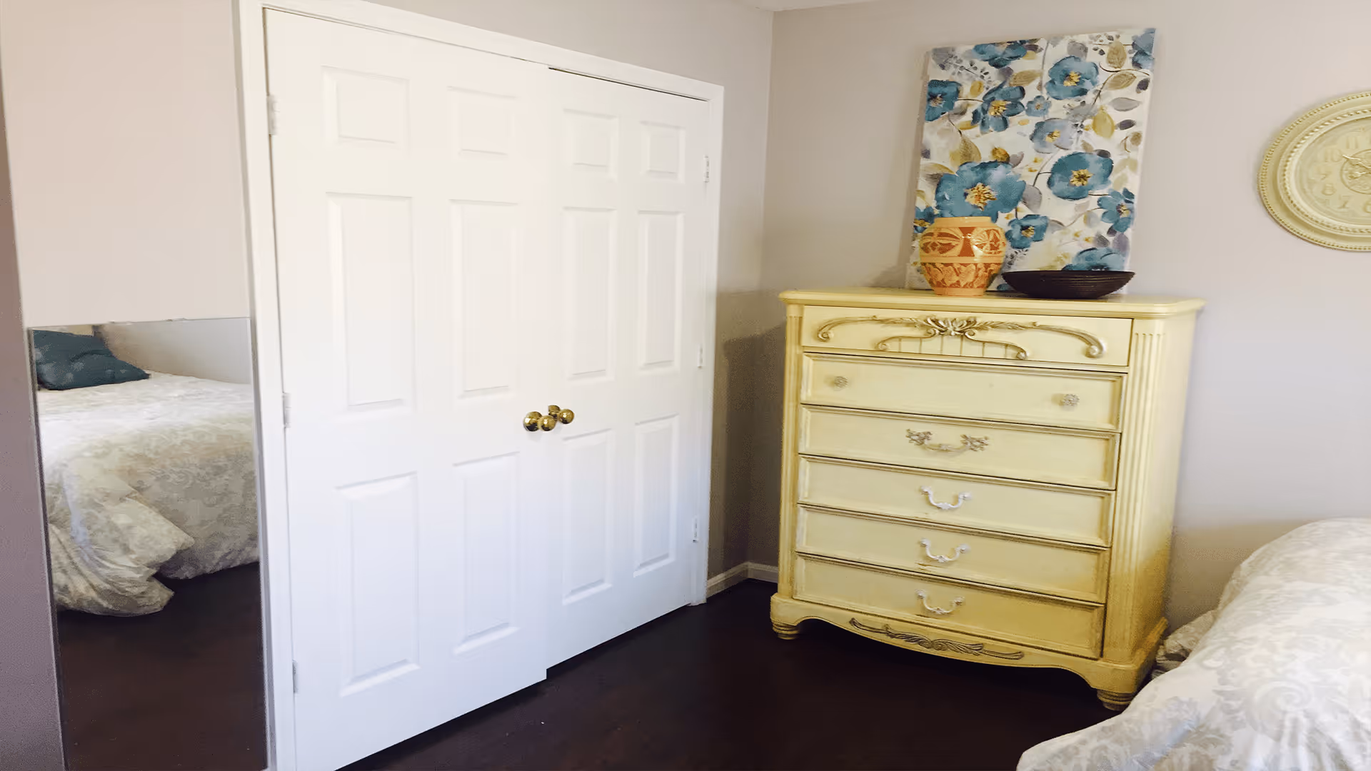 A bedroom with a white double-door closet, a yellow vintage-style dresser with decorative items on top, and a bed partially visible with a white patterned bedspread. A floral painting and a round wall decoration hang on the light-colored walls.